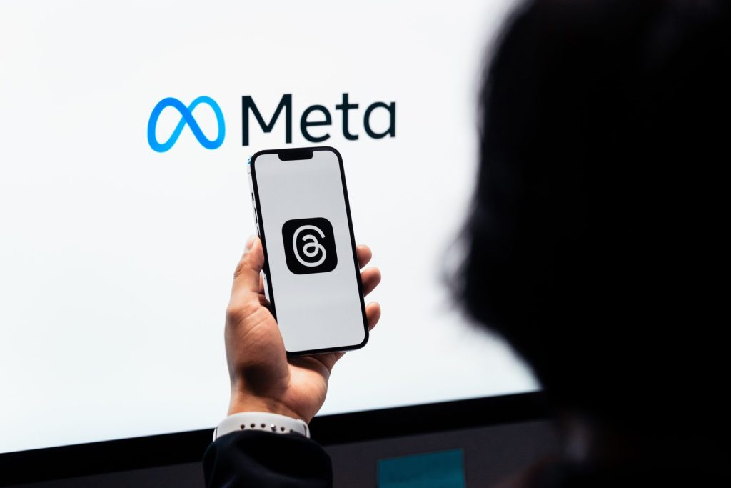 Person holding a smartphone displaying a logo with an @ in front of a screen showing the Meta logo.